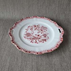 Pode incluir: Travessa de servir em cerâmica branca com um design floral vermelho e bordas onduladas. A travessa tem duas alças e uma borda decorativa. O centro apresenta um padrão floral e arquitetônico vermelho detalhado.