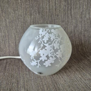 Op de afbeelding: Een bolvormige glazen lamp met een matte afwerking en een wit bloemenpatroon van takken en bloesems. De lamp heeft een brede ronde opening aan de bovenkant en is voorzien van een wit elektrisch snoer dat uit de zijkant van de voet komt.