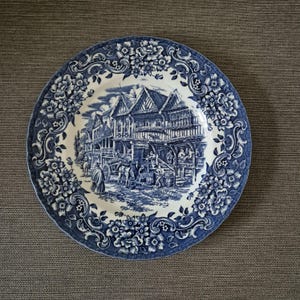 May include: A round ceramic plate featuring a blue and white transferware pattern. The border displays intricate floral motifs, while the center depicts a detailed scene of a historic street with timber-framed buildings, a horse-drawn cart, and figures.