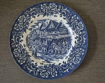May include: A round ceramic plate featuring a blue and white transferware pattern. The border displays intricate floral motifs, while the center depicts a detailed scene of a historic street with timber-framed buildings, a horse-drawn cart, and figures.