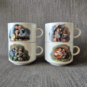 Könnte beinhalten: Vier weiße Keramikbecher, gestapelt in zwei Säulen, jeder mit einer eigenen Illustration im Vintage-Stil, die Kinder in verschiedenen Szenen im Freien zeigt. Die Becher haben einfache Henkel und eine glatte Oberfläche mit detaillierten Bildern.