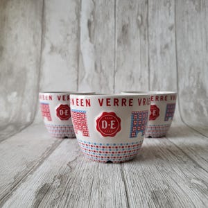 May include: Three white ceramic bowls with red and blue designs. The bowls have the text "Van Een Verre Vrie" and a red seal with the text "D.E. Anno 1752" on them.