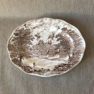Peut inclure: Assiette décorative marron et blanche avec un paysage. L'assiette représente un pont, des arbres et une personne en train de pêcher. Les bords de l'assiette ont un motif festonné.