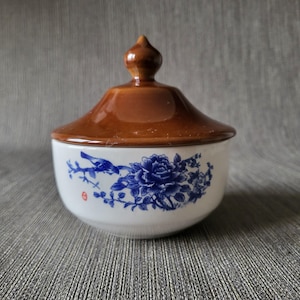 Könnte beinhalten: Eine weiße Keramik-Zuckerdose mit braunem Deckel. Die Schüssel hat ein blaues Blumen- und Vogeldesign. Der Deckel hat einen spitzen Knauf. Die Schüssel ist wahrscheinlich Vintage oder antik.
