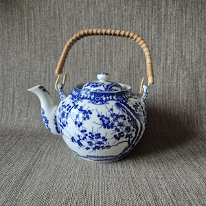 Op de afbeelding: Witte en blauwe keramische theepot met een bloemenpatroon. De theepot heeft een gevlochten handvat en een bijpassende deksel. De tuit en handvatbevestigingen zijn goudkleurig. Een klassiek ontwerp voor het serveren van thee.