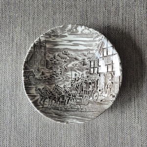 May include: A round, brown and white decorative plate with a detailed transferware design. The scene depicts a horse-drawn carriage, buildings, and figures. The plate has a glossy finish and is set against a gray background.