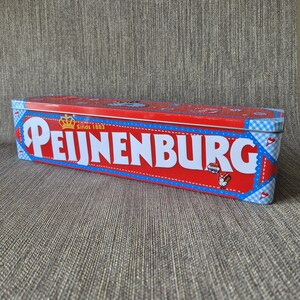 Op de afbeelding: Een rechthoekige rode blikken doos met het woord "PEIJNENBURG" in het wit, een kroon en de tekst "Sinds 1883". De doos heeft blauw-witte ruitjesaccenten en een cartoonafbeelding van een konijn.