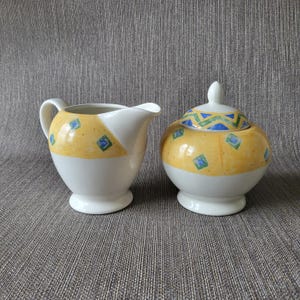 May include: A white ceramic creamer and sugar bowl set. The creamer has a handle and spout. The sugar bowl has a lid with a small knob. Both pieces have a yellow band with blue and green square designs.