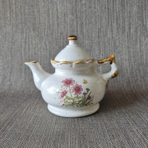 May include: A white ceramic teapot with a speckled finish, featuring a floral design of pink and purple flowers with a small yellow butterfly. The teapot has a scalloped rim, a brown-trimmed handle, and a matching lid with a brown knob.