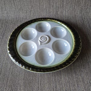 Könnte beinhalten: Ein Keramik-Schnecken-Teller mit sieben runden Vertiefungen. Der Teller ist hellblau mit einem dunkelgrünen und braunen Rand. In der Mitte des Tellers befindet sich ein Schneckenhaus-Design.