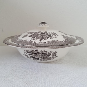 Puede incluir: Un cuenco de cerámica blanca con tapa, con un estampado floral en tonos grises. El cuenco tiene un borde ancho y un pomo decorativo en la tapa. El diseño floral incluye rosas y follaje, creando un aspecto clásico y elegante.