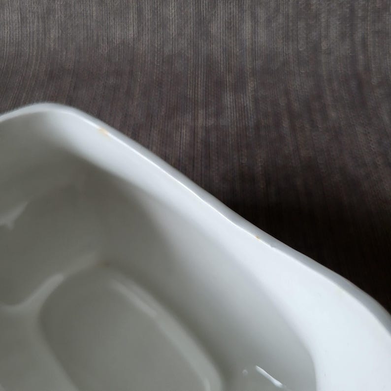 May include: A white, rectangular ceramic dish with gently curved edges. The dish has a glossy finish and appears to be empty. The background is a textured, dark gray fabric.