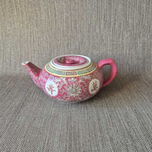 May include: A pink ceramic teapot with a matching lid and handle. The teapot features a floral design with Chinese characters and decorative trim in yellow, green, and white. The teapot is a traditional Asian design.
