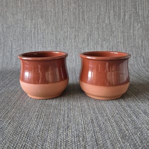 Op de afbeelding: Twee kleine, handgemaakte terracotta potten. De bovenste helft heeft een glanzend, roodbruin glazuur, terwijl de basis mat lichtbruin is. De potten hebben een afgeronde vorm en een eenvoudig ontwerp, perfect voor kleine planten of decoratie.