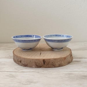 Könnte beinhalten: Zwei weiße Keramikschalen mit blauen dekorativen Mustern. Die Schalen haben eine abgerundete Form und stehen auf einer Holzscheibe. Die blauen Muster umfassen geometrische und wellenartige Designs. Die Schalen sind wahrscheinlich zum Servieren von Speisen gedacht.