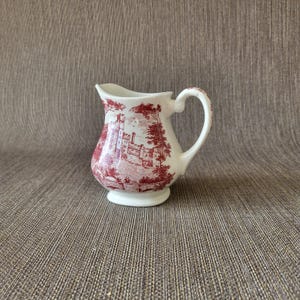 Op de afbeelding: Witte keramische kan met een rood transferware ontwerp van een kasteel en bomen. De kan heeft een gebogen handvat en een uitlopende tuit. De basis is een kleine, afgeronde voet.