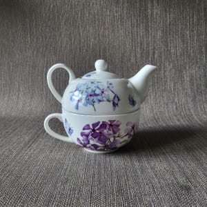 Könnte beinhalten: Weißes Keramik-Tee-für-eins-Set mit einem Blumendesign in Blau- und Lilatönen. Das Set beinhaltet eine Teekanne mit Deckel und eine Teetasse. Die Teekanne steht auf der Teetasse. Das Set steht auf einer grau strukturierten Oberfläche.