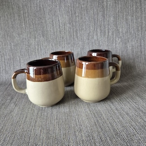 May include: A set of four ceramic mugs featuring a two-tone design with a dark brown upper section and a cream-coloured lower section. Each mug has a rounded body, a smooth glossy finish, and a sturdy handle attached to the side for easy gripping.