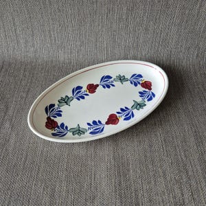 May include: Oval serving platter with a white base and a decorative floral border. The border features blue, red, and green leaves and flowers. A thin red line encircles the edge of the platter.