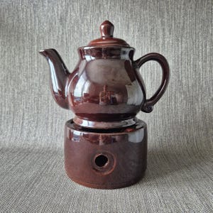 Peut inclure: Une théière en céramique brun foncé brillant avec un couvercle et une anse assortis, posée sur un socle chauffe-thé cylindrique en céramique. La base présente une ouverture circulaire sur le devant pour accueillir une source de chaleur.