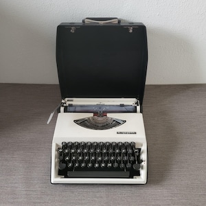 Puede incluir: Una máquina de escribir Adler Tippa vintage en un estuche de transporte negro. La máquina de escribir es blanca con teclas negras y un carro cromado. El estuche está abierto, revelando la máquina de escribir lista para usar. La palabra "Tippa" está impresa en la parte delantera.