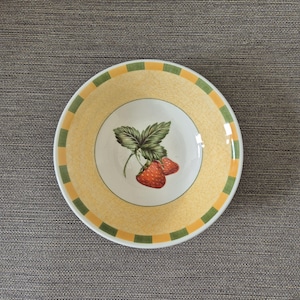Puede incluir: Un cuenco redondo de cerámica con borde amarillo y verde. El centro del cuenco presenta un círculo blanco con una ilustración detallada de dos fresas rojas y hojas verdes. El cuenco tiene un fondo amarillo texturizado.