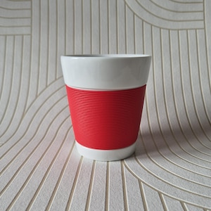 Op de afbeelding: Een witte keramische beker met een brede, felrode siliconen hoes met een horizontale geribbelde textuur. De beker heeft een gladde, glanzende afwerking aan de boven- en onderkant, wat een contrast vormt met de matte, getextureerde grip.