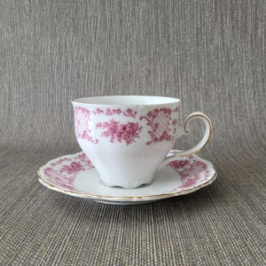 Può includere: Servizio da tè bianco con motivo floreale rosa. La tazza ha manico e bordo dorati, e il piattino presenta un motivo floreale abbinato e bordatura dorata. Il set è su sfondo grigio.