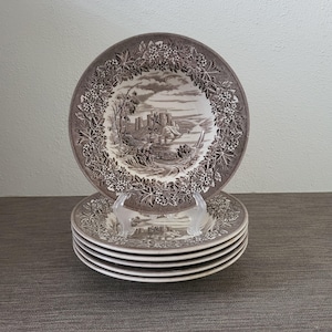 May include: A set of six brown and white ceramic plates. Each plate features a detailed landscape scene with a castle and a floral border. The plates are stacked on a gray surface.