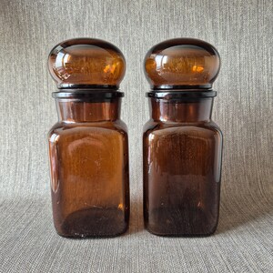 Op de afbeelding: Twee amberkleurige glazen apothekerspotten met afgeronde, donkerbruine stoppers. De potten hebben een rechthoekige vorm en worden tegen een neutrale achtergrond gepresenteerd. De potten worden waarschijnlijk gebruikt voor het bewaren van droge waren of andere items.