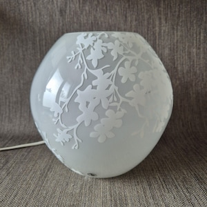 Peut inclure: Lampe en verre dépoli avec un motif floral. La lampe est de couleur blanc doux et de forme arrondie. Des motifs floraux blancs sont gravés sur la surface, créant une esthétique délicate et élégante. Un cordon blanc s'étend de la base.