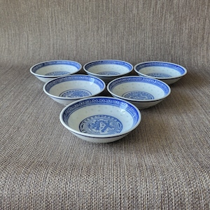 May include: Six white and blue porcelain bowls with a traditional Chinese design. Each bowl features a blue rim and a central blue motif, with a delicate dotted pattern around the bowl's interior. The bowls are arranged on a textured brown surface.