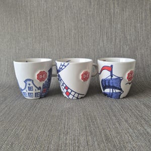 Op de afbeelding: Drie witte keramische mokken met blauwe en rode illustraties van een Nederlands huis, een windmolen en een zeilschip. Elke mok heeft een rood, cirkelvormig reliëf-embleem aan de zijkant met de witte tekst D.E 1753 op het oppervlak.
