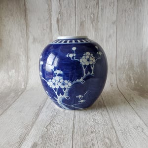 Puede incluir: Un jarrón de porcelana azul y blanco con un diseño floral. El jarrón es redondo y tiene una boca ancha. Está decorado con flores blancas y ramas sobre un fondo azul cobalto.