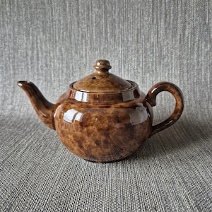 May include: A brown ceramic teapot with a lid, spout, and handle. The teapot has a glossy finish and a speckled pattern. The handle is curved, and the spout is slender. The lid has a small knob on top. The teapot is designed for brewing tea.