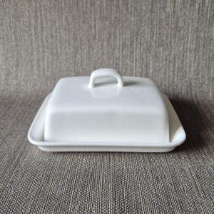 Könnte beinhalten: Eine weiße Butterdose aus Keramik mit einem rechteckigen Unterteil mit leicht erhöhtem Rand und einem passenden rechteckigen Deckel. Der Deckel verfügt über einen mittig platzierten, gewölbten Griff auf der Oberseite.