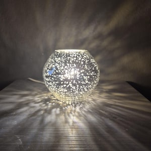 Könnte beinhalten: Eine kugelförmige Glaslampe mit silbernem Quecksilber-Finish und einem gesprenkelten Sternenmuster. Die Lampe strahlt ein warmes Licht von innen aus und wirft komplexe, strahlenförmige Schatten von ihrem Sockel aus in die Umgebung.