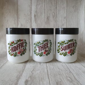 May include: Three white ceramic canisters with black lids and green and red floral designs. The canisters are labeled "Koffie", "Thee", and "Suiker".
