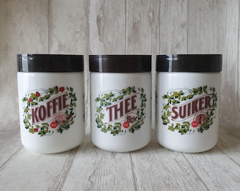 May include: Three white ceramic canisters with black lids and green and red floral designs. The canisters are labeled "Koffie", "Thee", and "Suiker".