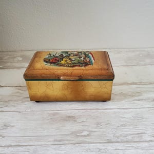Op de afbeelding: Een vintage houten doos met een geschilderd tafereel van mensen in een boot. De doos heeft een verweerde gouden afwerking en een groene rand.