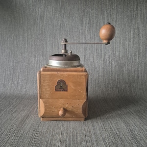 Op de afbeelding: Een vintage houten koffiemolen met een metalen bovenkant en een slinger. De molen heeft een kleine lade en een houten knop. Een metalen plaatje met tekst is aan de voorkant bevestigd. Het hout heeft een lichtbruine kleur.