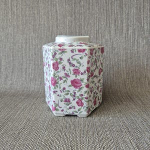 Puede incluir: Un tarro de cerámica hexagonal con base blanca decorado con un patrón repetitivo de rosas rosadas, pequeñas flores moradas y hojas verdes. El recipiente tiene una abertura superior plana y un acabado liso y brillante en sus seis lados.
