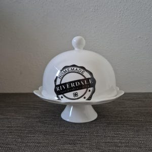 May include: White ceramic cloche with a round lid and a pedestal base. The lid features a black and white emblem that reads "Homemade Riverdale fashionable living". The cloche is designed for serving and displaying food.