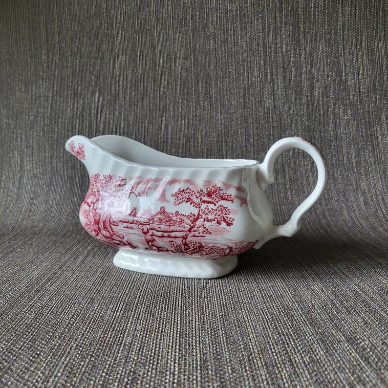 May include: White ceramic gravy boat with a red pastoral scene design. The boat has a curved spout, a handle, and a pedestal base. The red design features trees, buildings, and figures.