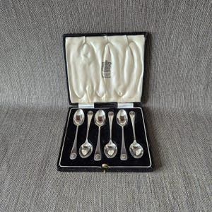 May include: A set of six silver spoons presented in a black velvet box with a cream satin lining. Each spoon features a shell design on the handle. The box is open, displaying the spoons and a crest.