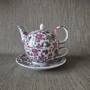 Könnte beinhalten: Ein floral gemustertes Teeservice mit Teekanne, Tasse und Untertasse. Das Set hat einen weißen Grund mit einem Design aus rosa, lila und grünen Blumen. Die Teekanne hat eine runde Form mit Henkel und Ausguss. Die Tasse passt in die Teekanne.