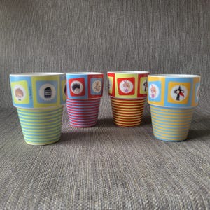 Op de afbeelding: Vier keramische bekers met kleurrijke ontwerpen. Elke beker heeft een gestreepte basis in verschillende kleuren: geel en blauw, rood en blauw en geel en rood. De bovenkant van elke beker heeft een vierkant ontwerp met cartoon illustraties en de tekst "Lum".