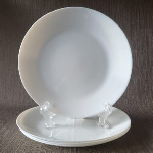 May include: A stack of white, round plates. The plates are made of a glossy material and are displayed on clear plastic plate stands. The plates have a simple, clean design, suitable for various dining settings.