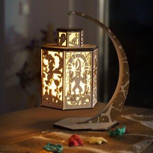 Könnte beinhalten: Eine dekorative Holzlaterne mit Halloween-Motiv. Die Laterne zeigt ausgeschnittene Silhouetten von Geistern, Werwölfen und anderen gruseligen Figuren. Sie wird von innen beleuchtet und an einem gebogenen Ständer aufgehängt. Die Laterne steht auf einer Holzoberfläche.