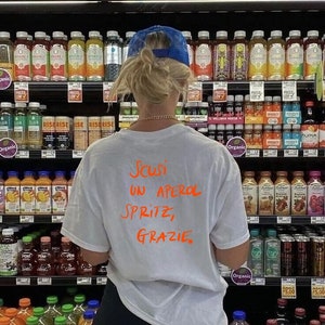 May include: A white t-shirt with the text "Scusi un Aperol Spritz, Grazie." written in orange marker.